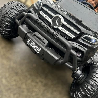 A black Mercedes-Benz branded kids' ride-on toy truck with large off-road tires, shown with a child sitting inside. The toy features a chrome-style front grille, a black front bumper with a 'L3GHTN' brand plate, and a wet surface beneath it.